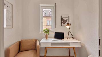 Una habitación pequeña y bien iluminada con un escritorio blanco, una laptop, una planta en maceta, una lámpara y un cuadro en la pared.