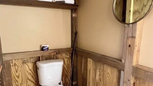 A small, wooden-paneled bathroom with a toilet, a shelf above the toilet holding towels, and a mirror on the wall.
