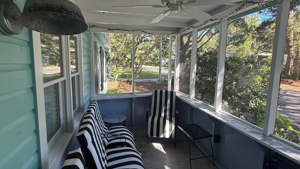 Uma varanda coberta com ventilador de teto, um banco listrado, uma mesa pequena e janelas amplas com vista para o exterior.