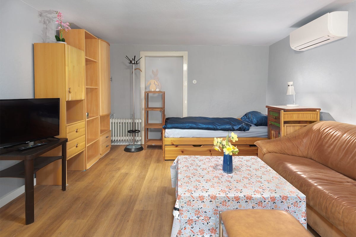 A compact room with a bed, a television, a wooden wardrobe, a small table with a floral tablecloth, and a brown leather sofa.