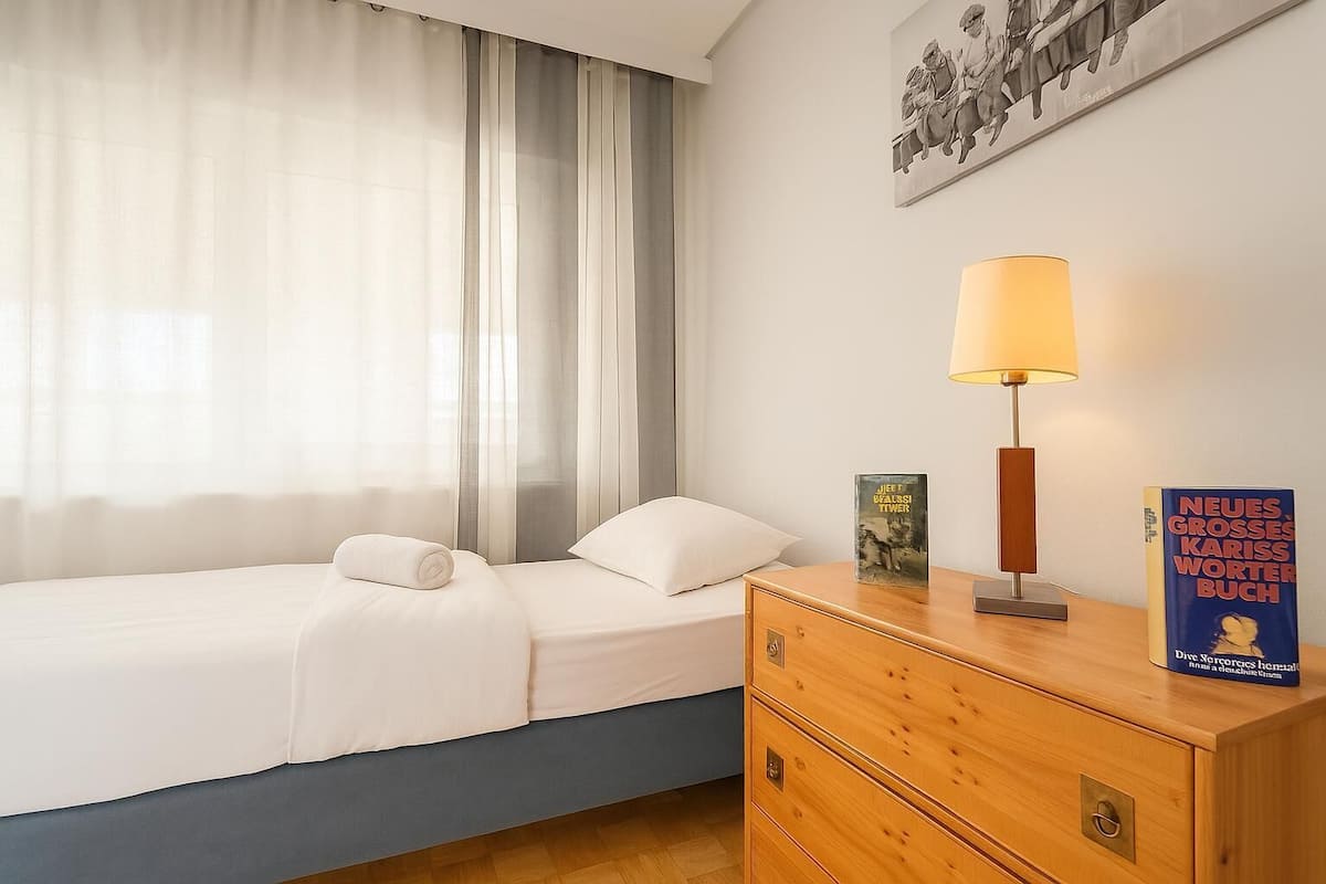 A bedroom with a bed, a bedside table with a lamp, a book, and a wall-mounted painting.