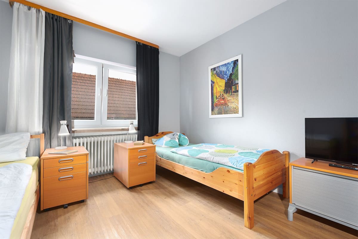 A bedroom with a bed, a desk, a radiator, a window with curtains, and a painting on the wall.