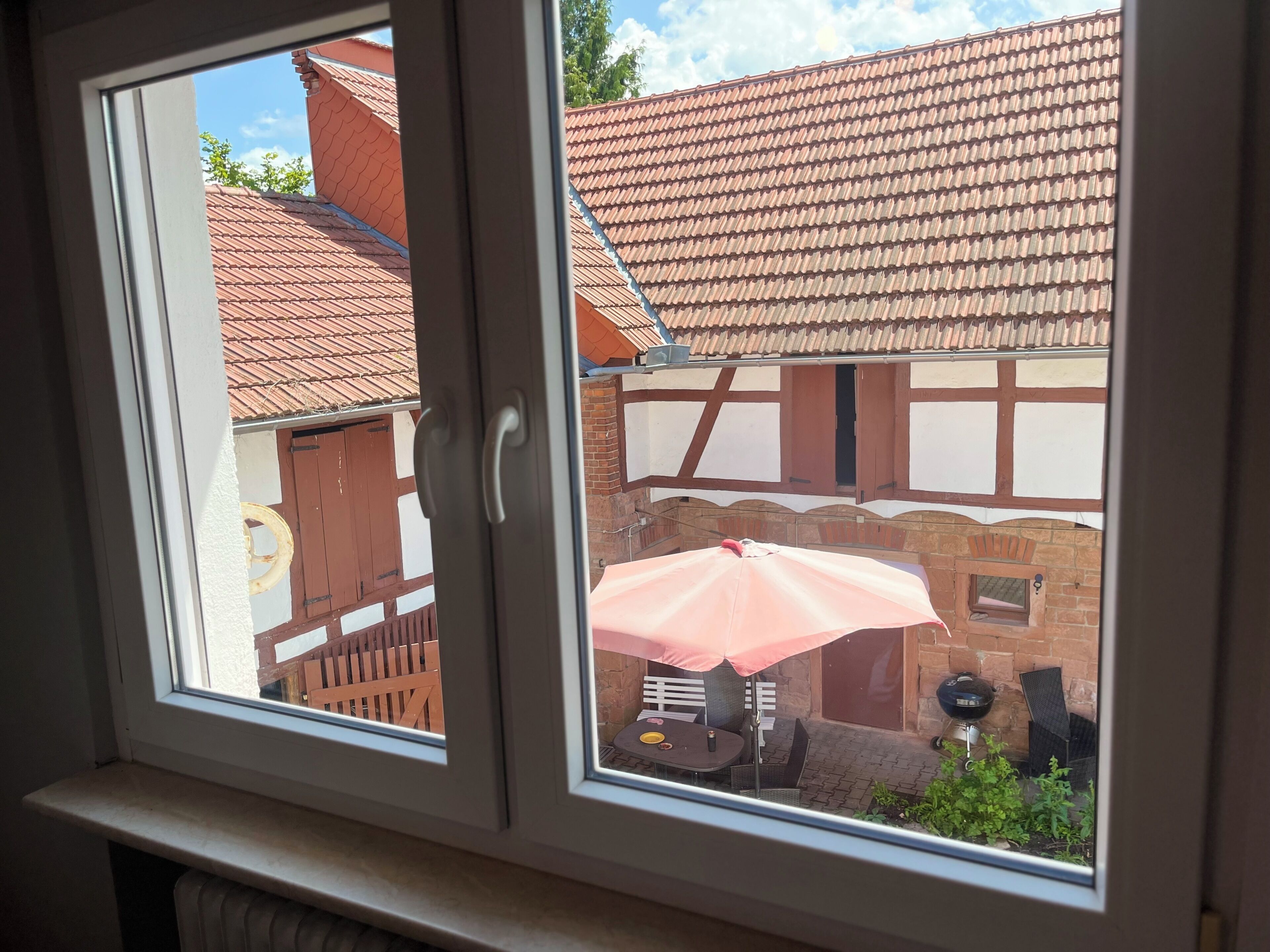 A window with a view of a courtyard featuring a brick building, a wooden fence, and a table with an umbrella.
