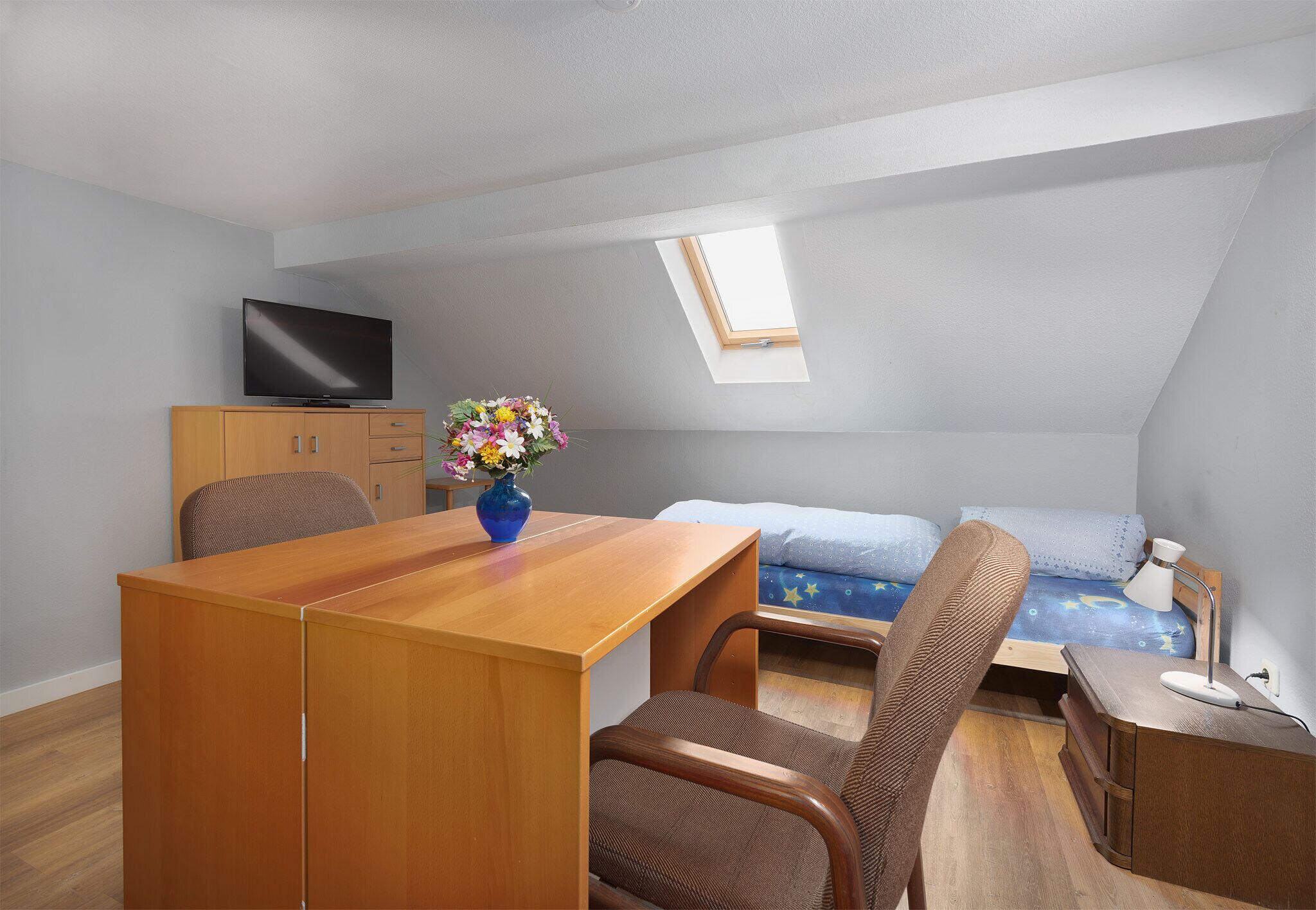 Quarto compacto com escrivaninha de madeira, uma cadeira, cama com colcha azul, mesa de cabeceira pequena com luminária, televisão em um armário e um claraboia.