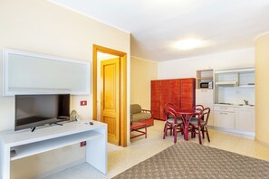 A compact living space with a TV on a white stand, a dining area with red chairs, a small kitchen with a microwave, and a red cabinet. - Charming Rina Sea Studio Apartment-central Air-con & Wifi Sleeps 2 Persons (Alghero)
