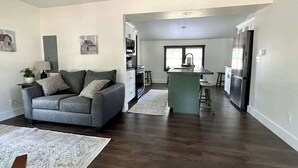 A modern living room with a grey sofa, a kitchen area with a fridge, and a dining table. - Lake Street Retreat - Walk to the beach! (Central Lake)