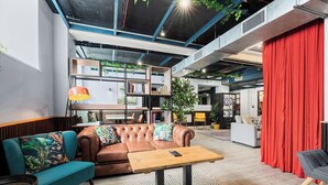 Un espacio lounge moderno con un sofá de cuero, un sillón color turquesa y una mesa de centro de madera. Se observan estanterías, una máquina expendedora y una cortina roja al fondo.
