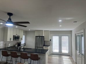 A modern kitchen with white cabinets, a stainless steel refrigerator, a black countertop island with bar stools, and a ceiling fan. - "Abigail", Fenced, Dog friendly Single Level home, in Charming Downtown Apex (Apex)