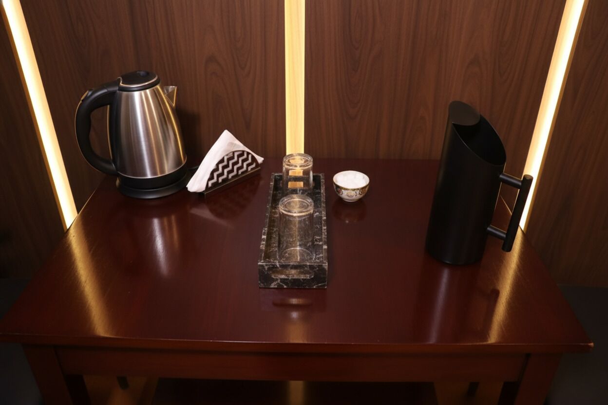 A wooden table with a stainless steel kettle, a napkin, a glass, and a black pitcher.
