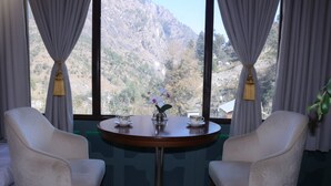 A room with two white armchairs, a round wooden table with a vase of flowers, and a view of mountains through large windows with grey curtains. - SnugBug Kasol (Kasol)
