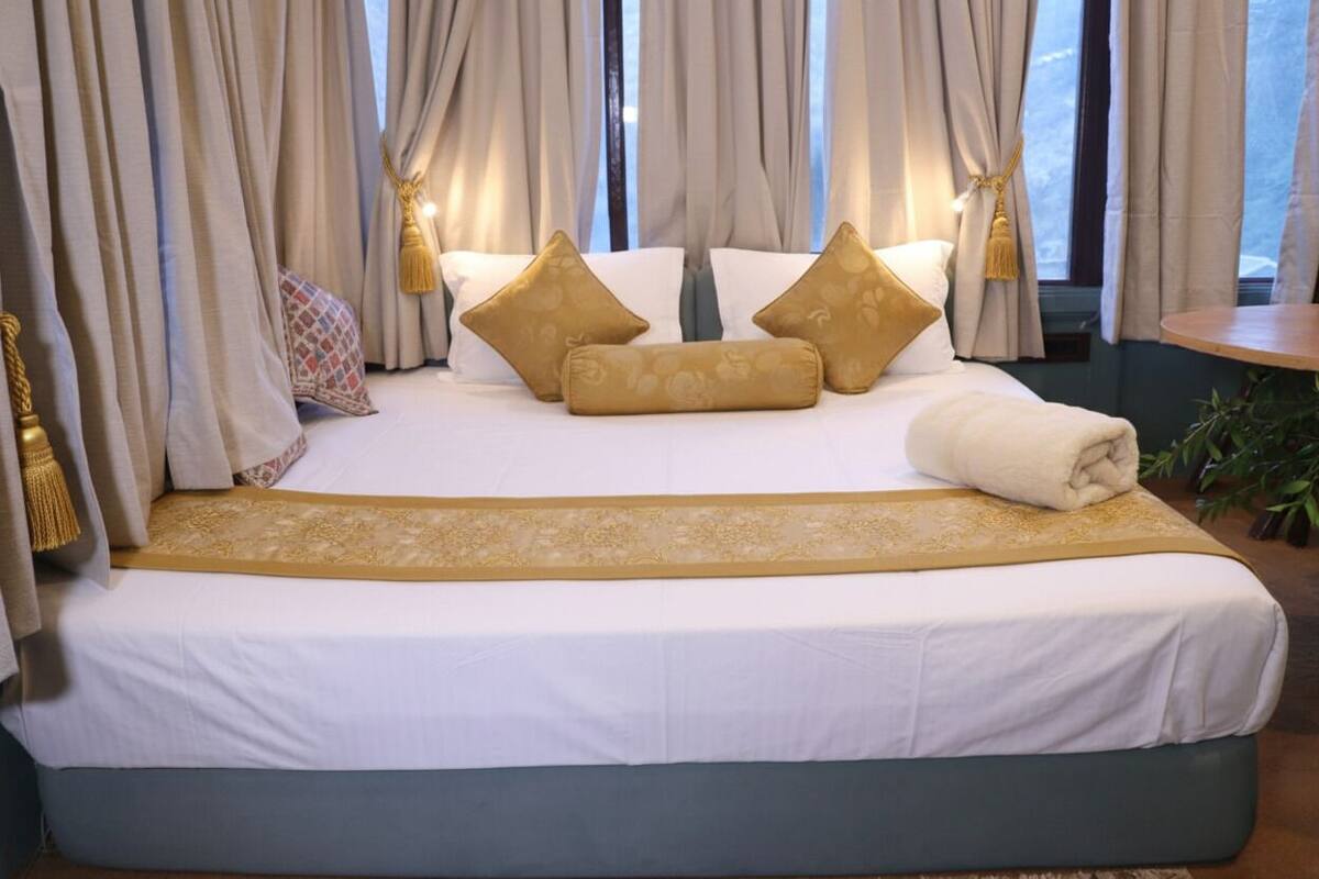 A neatly made bed with white and gold bedding, a decorative headboard, and a view of the outdoors through large windows.