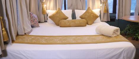 A neatly made bed with white and gold bedding, a decorative headboard, and a view of the outdoors through large windows.