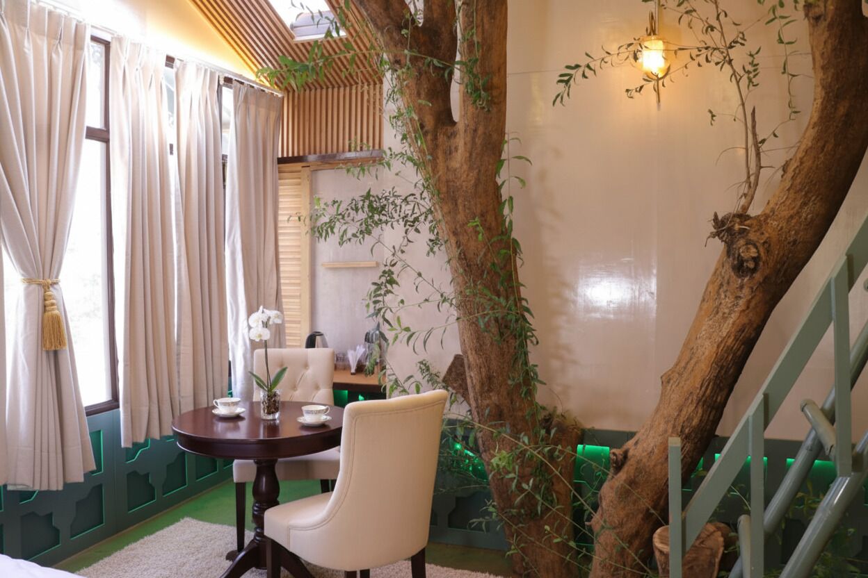 A dining area with a round table, white chairs, and a vase with an orchid. A large tree trunk is positioned to the right of the table, and a staircase with green railings is visible in the background.