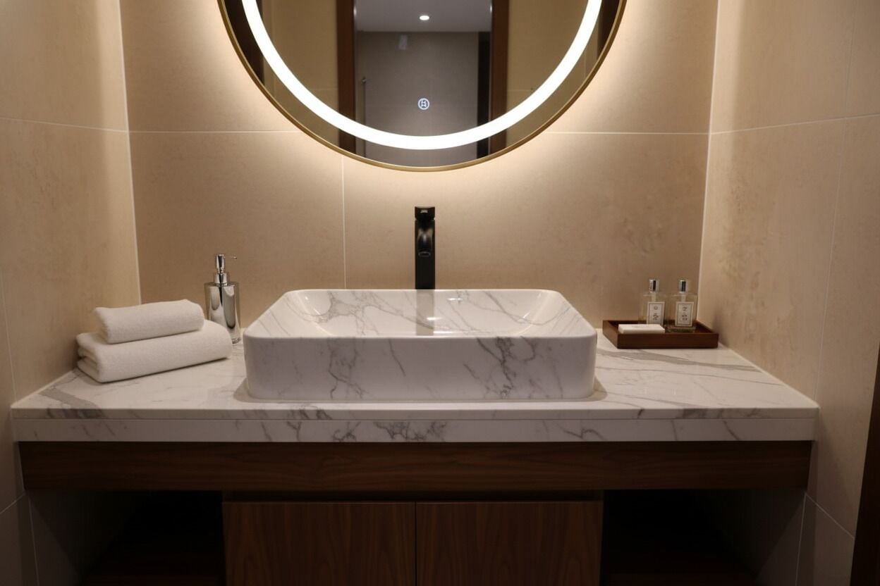 Un baño moderno con un lavabo de mármol, un espejo redondo con luz y un mueble de tocador de madera.