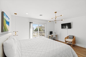 Une chambre moderne avec un grand lit, un bureau, une chaise, une télévision et un balcon offrant une vue sur les bâtiments et la verdure.