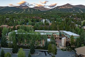 Zona residencial amb cases envoltades per un bosc dens, un edifici gran amb teulada verda i muntanyes al fons.