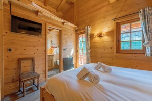 Ein Zimmer in einer Holzhütte mit einem Bett, einem Sessel, einem Fernseher und einem Fenster mit Vorhängen.