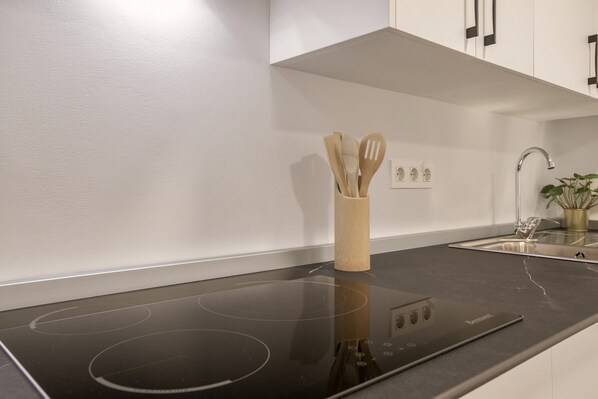 A modern kitchen with a black induction cooktop, wooden utensils in a holder, a stainless steel sink, and a potted plant.