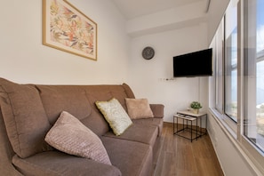 A modern living room with a brown sofa, a small side table with a plant, a mounted TV, and a large window.
