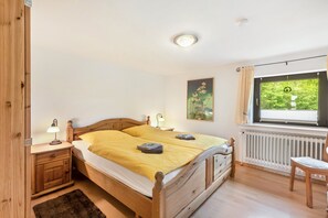Un lit en bois recouvert d’une couverture jaune, une table de chevet avec une lampe, une fenêtre donnant sur la verdure et un radiateur.