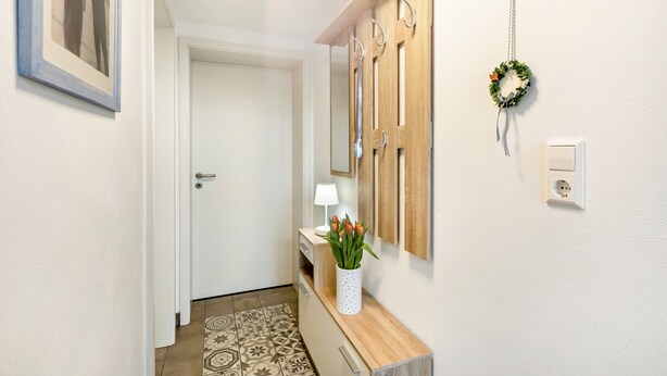 Un couloir avec une console en bois, un vase blanc contenant des fleurs, un miroir fixé au mur, une porte et une prise électrique.