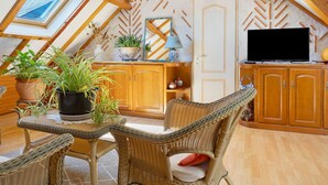 Une pièce mansardée chaleureuse avec un plafond en pente, des poutres apparentes, une table à manger entourée de chaises en osier, un téléviseur et des plantes en pot.
