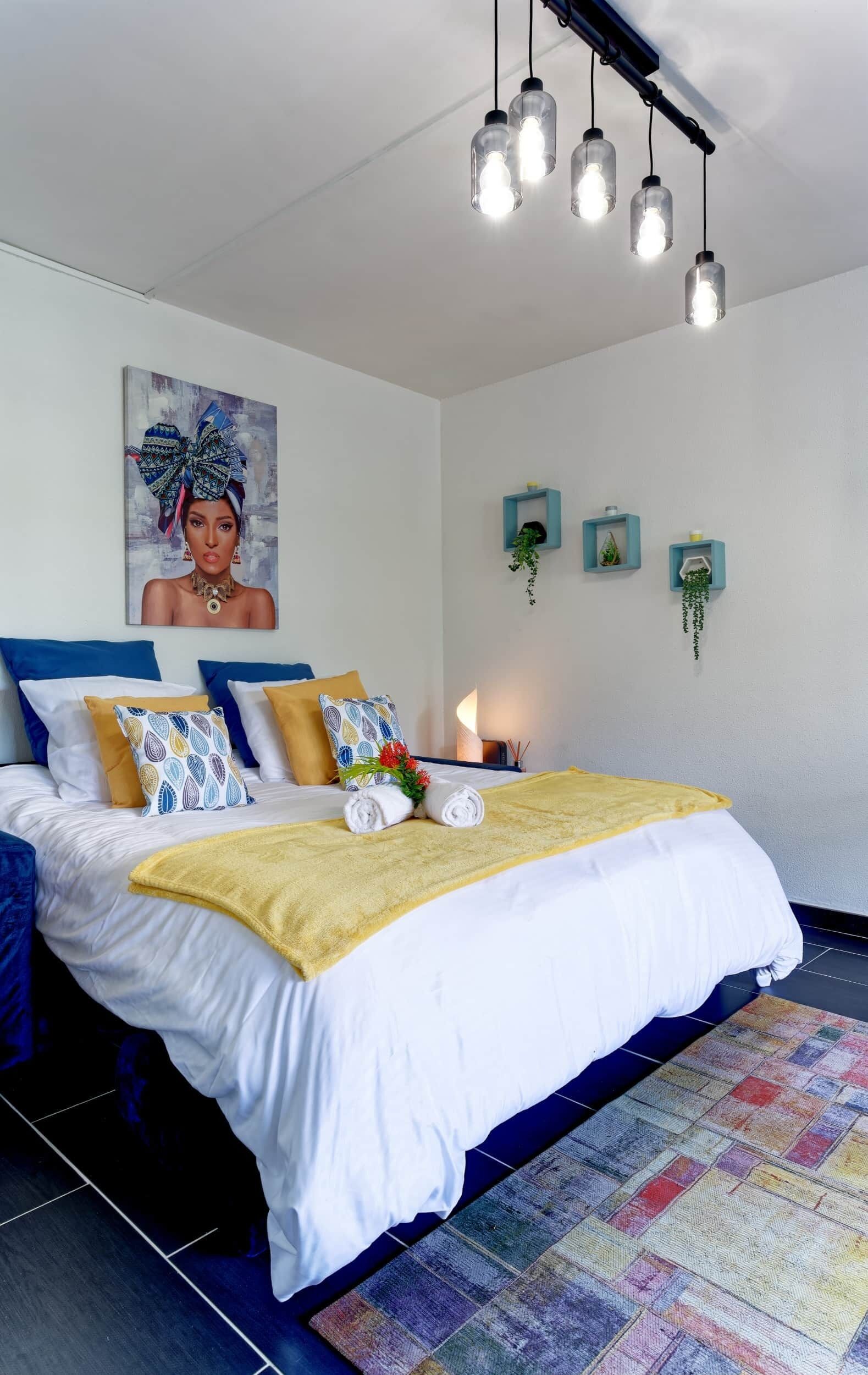 A neatly made bed with white and yellow bedding, a colorful rug, and a painting on the wall.