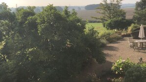 Une vue d’un paysage rural avec un chemin de terre, des arbres, une table, des chaises et un parasol.