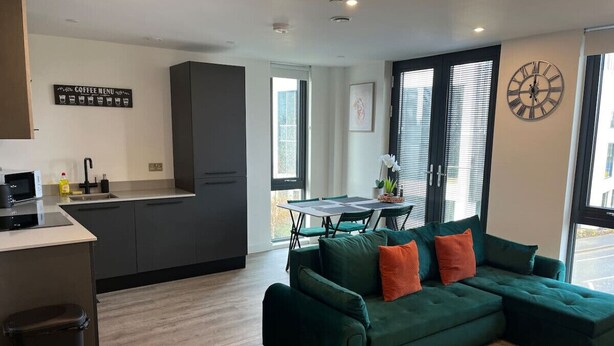 Een moderne keuken met donkere kasten, een eethoek met een tafel en stoelen, en een groene modulaire bank met oranje kussens.