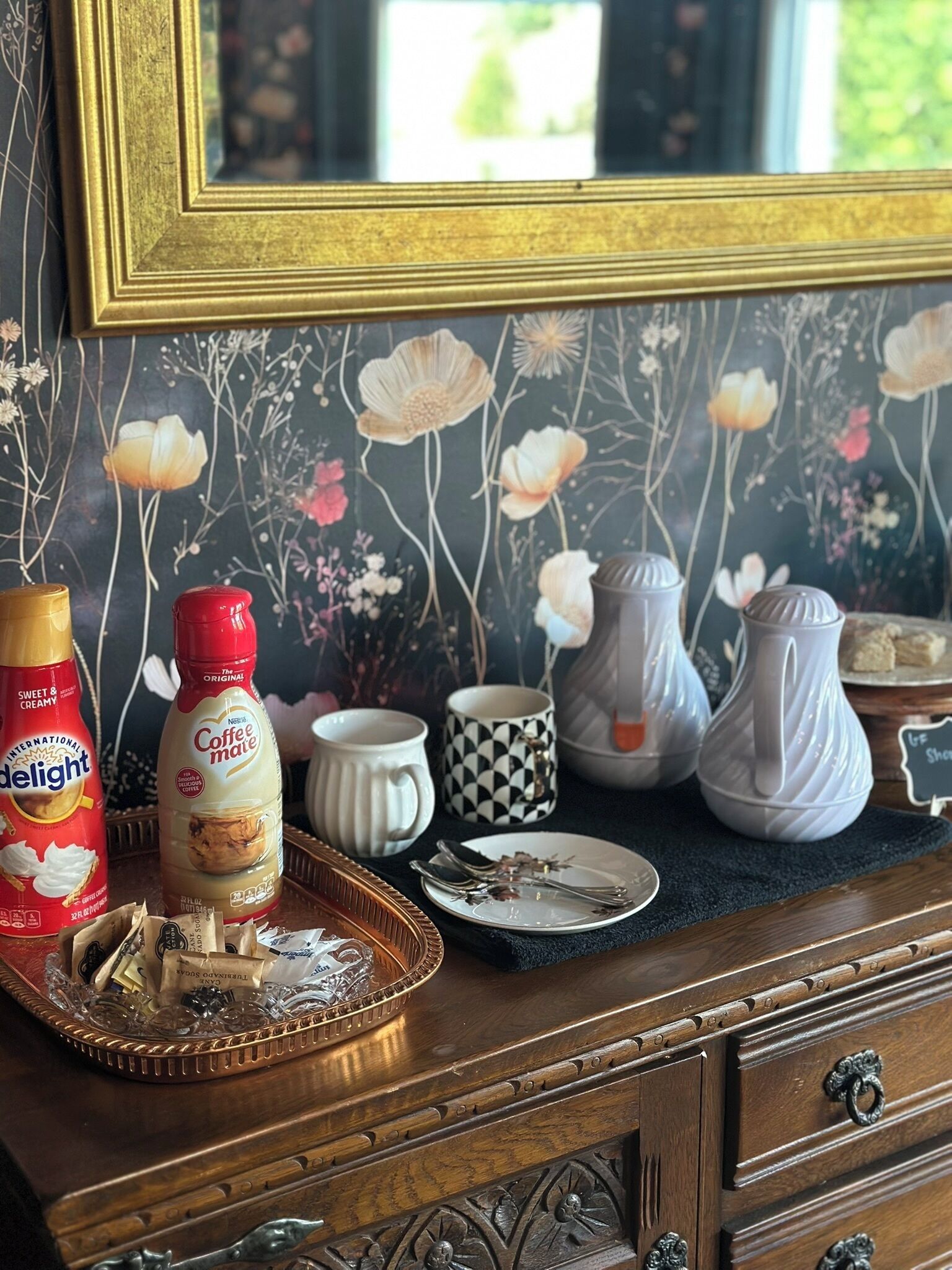 Un aparador de madera con un fondo de papel tapiz floral, una botella de café, una bandeja con bolsitas de té y un plato pequeño con cubiertos.