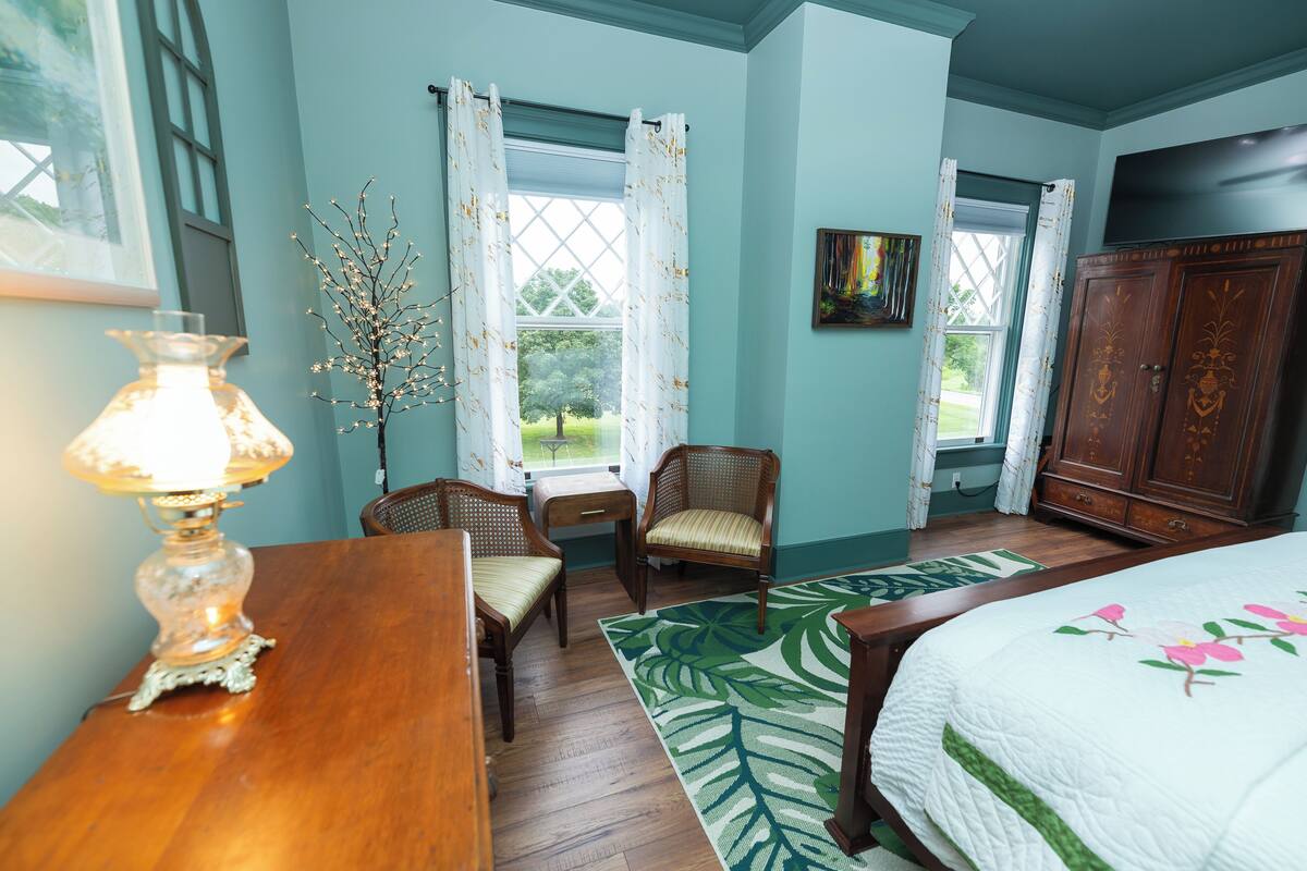 A bedroom with a bed, a wooden dresser, a desk, two chairs, a lamp, a window with curtains, and a painting on the wall.