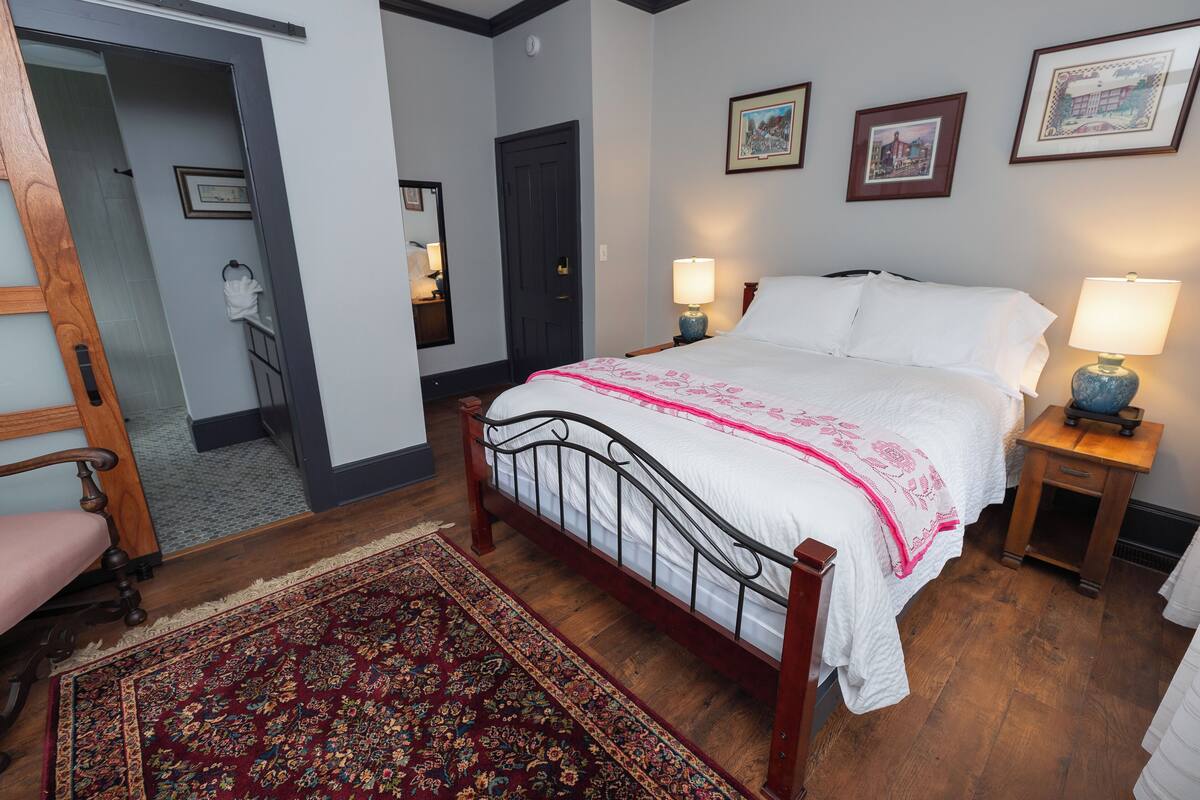 A bedroom with a wooden bed, two bedside lamps, a nightstand, a chair, a door, and a rug.