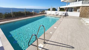 Une piscine aux eaux cristallines, avec un escalier en métal, des chaises longues blanches et un mur en pierre.