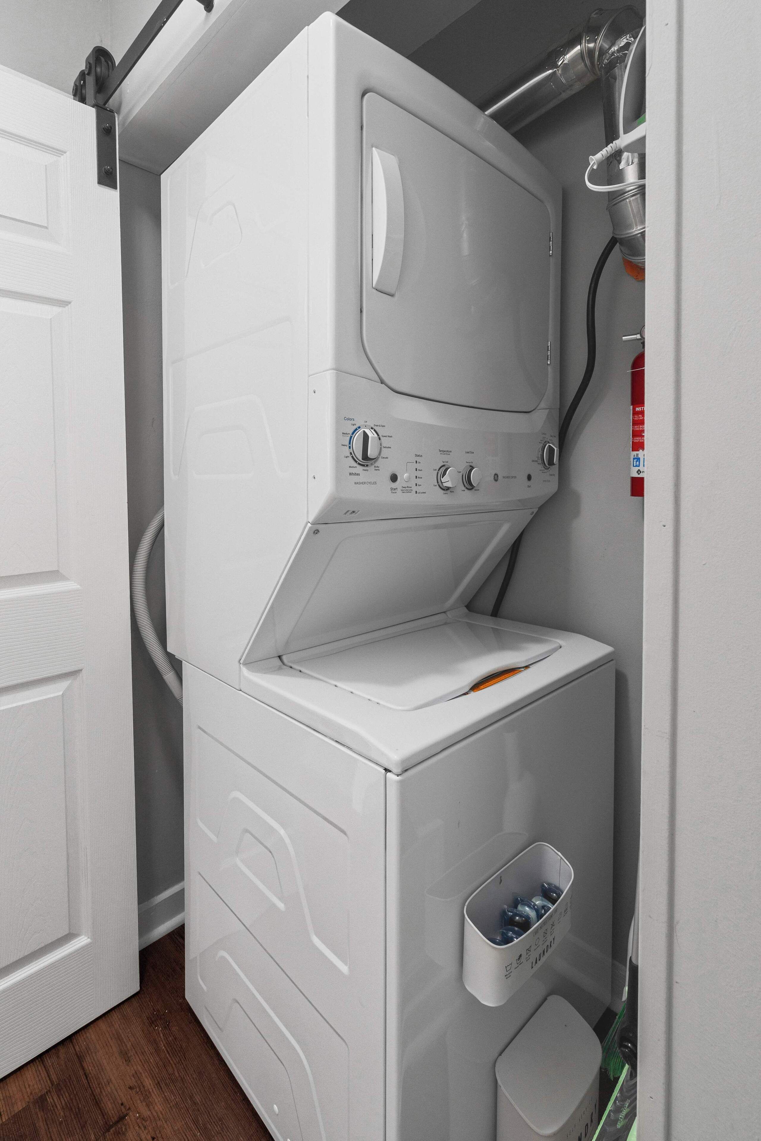 A compact, white laundry room with a front-loading washer and dryer, a small storage bin, and a hose for water connection.