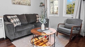 Una sala de estar moderna con un sofá gris, una mesa de centro de madera y un sillón gris. Hay una ventana con cortinas, un cuadro en la pared y una planta pequeña en el sofá.