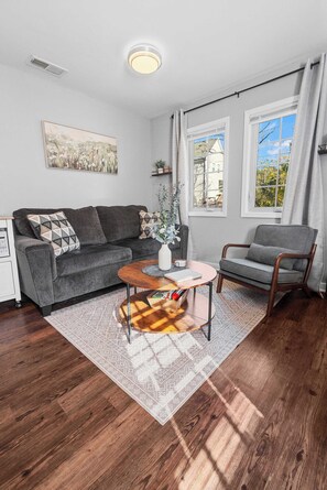 A modern living room with a grey sofa, a wooden coffee table, and a grey armchair. There is a window with curtains, a painting on the wall, and a small plant on the sofa. - 3 Min to Downtown | 7 to Duke | Washer + Dryer (Durham)