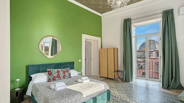 A bedroom with a green wall, a bed with patterned bedding, a round mirror, a wardrobe, and a view of buildings through a window with green curtains. - Antica Dimora Deluxe with Balcony, Wi-Fi and Air Conditioning (Napoli)