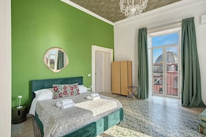 A bedroom with a green wall, a bed with patterned bedding, a round mirror, a wardrobe, and a view of buildings through a window with green curtains.
