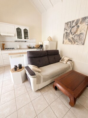 A modern living room with a sofa, a coffee table, and a kitchen area in the background. - Villa Ti Gîte 3*** Bougainvillier (Le Marin)