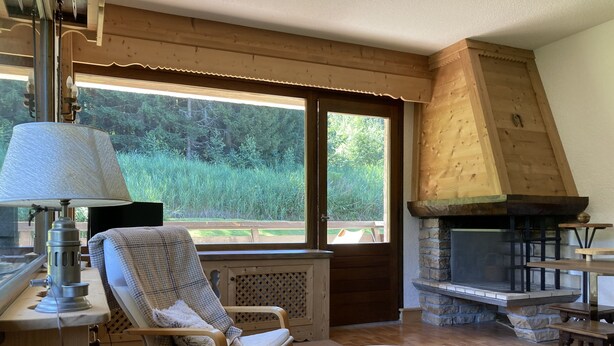 Ein gemütliches Zimmer mit Kamin aus Stein, einem großen Fenster mit Blick ins Freie, einem bequemen Sessel und einem Tisch mit einer Metallschale.