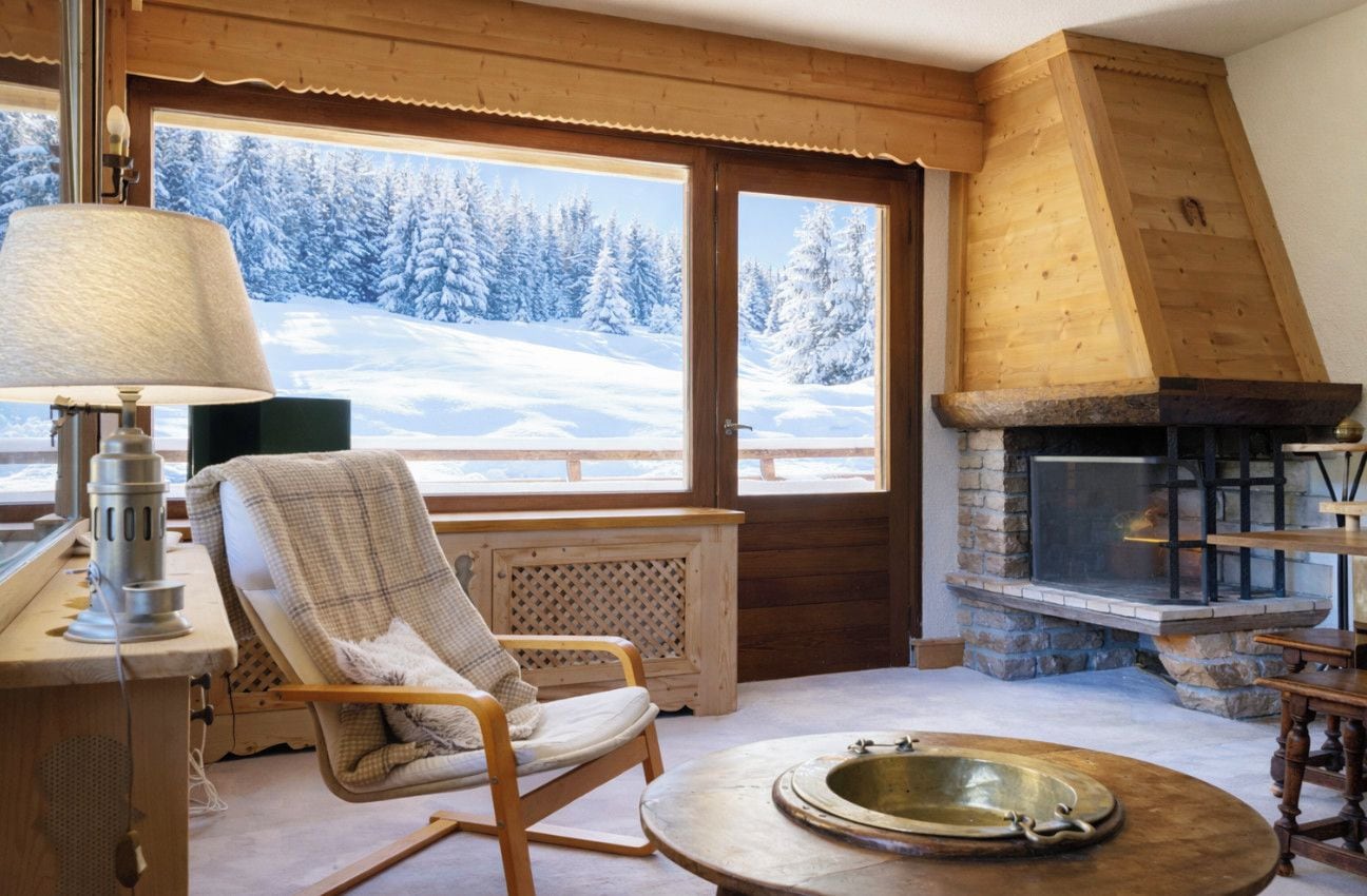 A cozy room with a stone fireplace, a wooden chair, a table with a brass tray, and a view of a snowy landscape through large windows.
