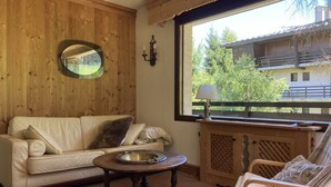 Ein gemütliches Zimmer mit einer Holzwand, einem runden Spiegel, einer Couch, einem kleinen runden Tisch, einem Sessel und einem Fenster mit Blick auf ein Gebäude und Bäume.