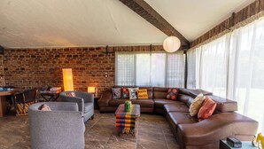 Een gezellige woonkamer met een metselwerk muur, een bruine leren bank, een grijze fauteuil en een kleurrijk geweven poef.