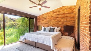Slaapkamer met een bakstenen muur, een plafondventilator, een bed met wit beddengoed en uitzicht op groen buiten.