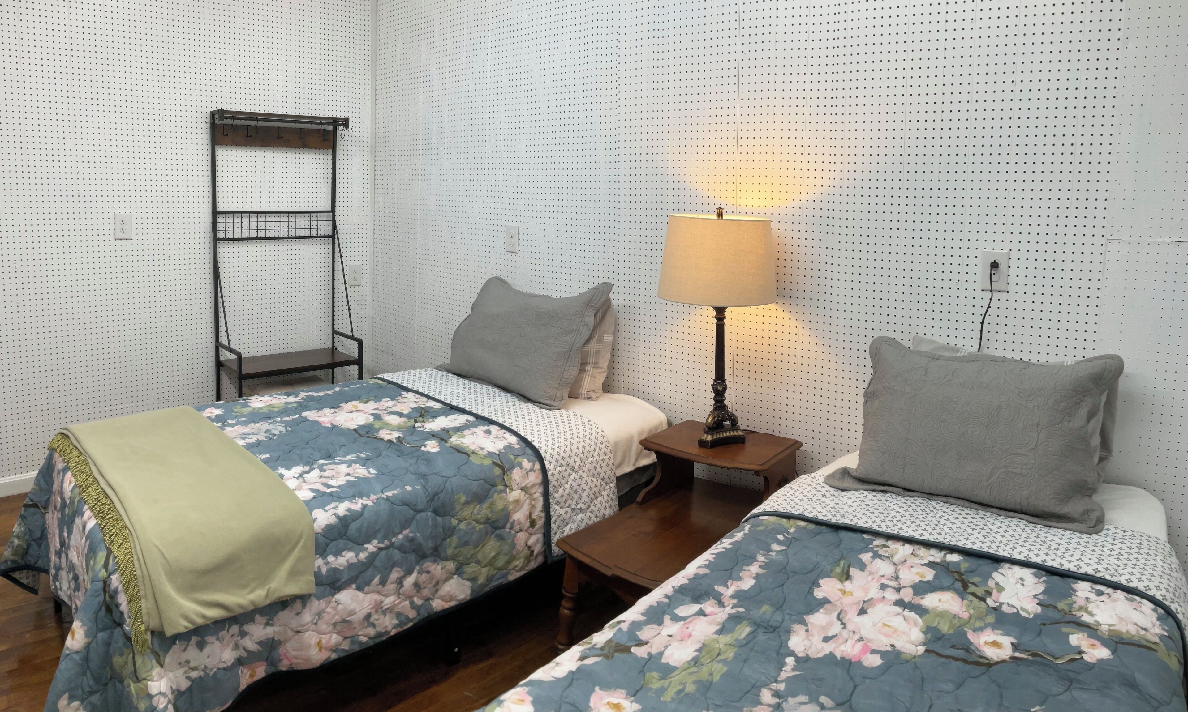 Une chambre avec deux lits simples, une table de chevet avec une lampe, une étagère fixée au mur et une prise de courant.
