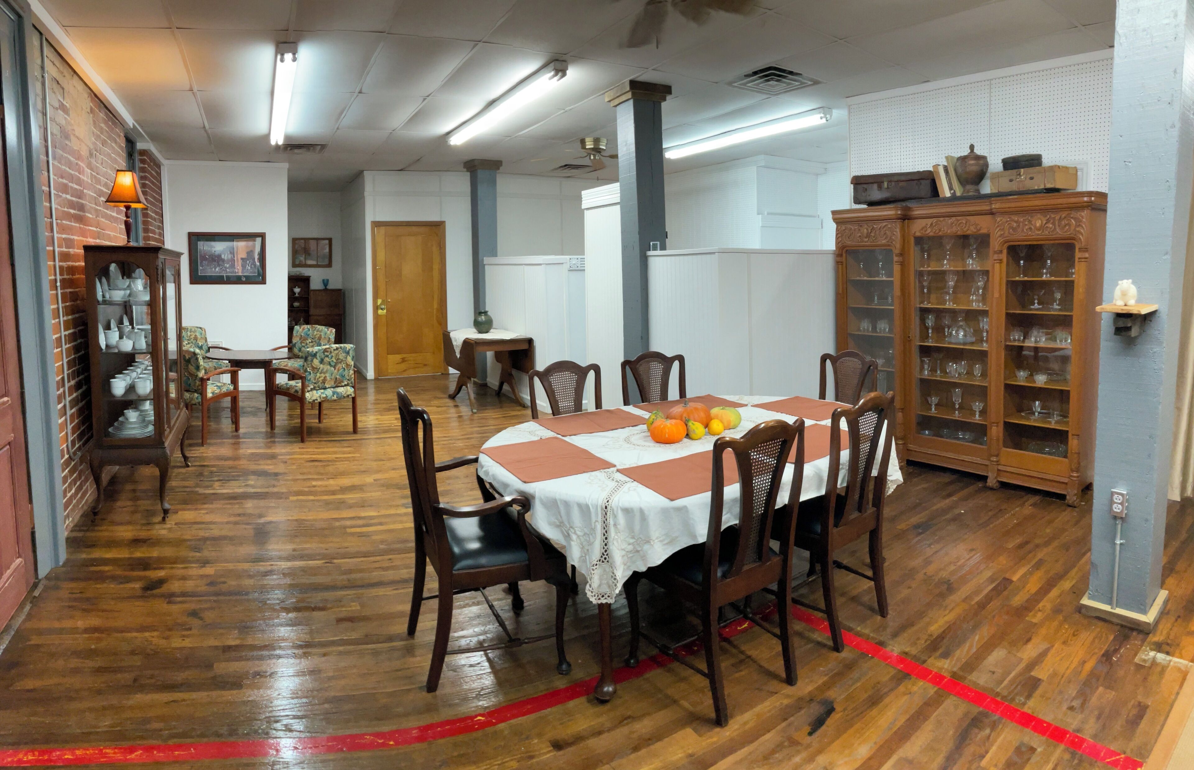 Un coin repas avec une table ronde dressée pour six personnes, entourée de chaises en bois. Un buffet en bois avec des portes en verre se trouve à droite, et une étagère murale ornée d’objets décoratifs est visible.