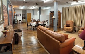 Um quarto espaçoso com piso de madeira, um sofá de couro marrom, uma mesa de jantar com cadeiras e uma estante de livros.