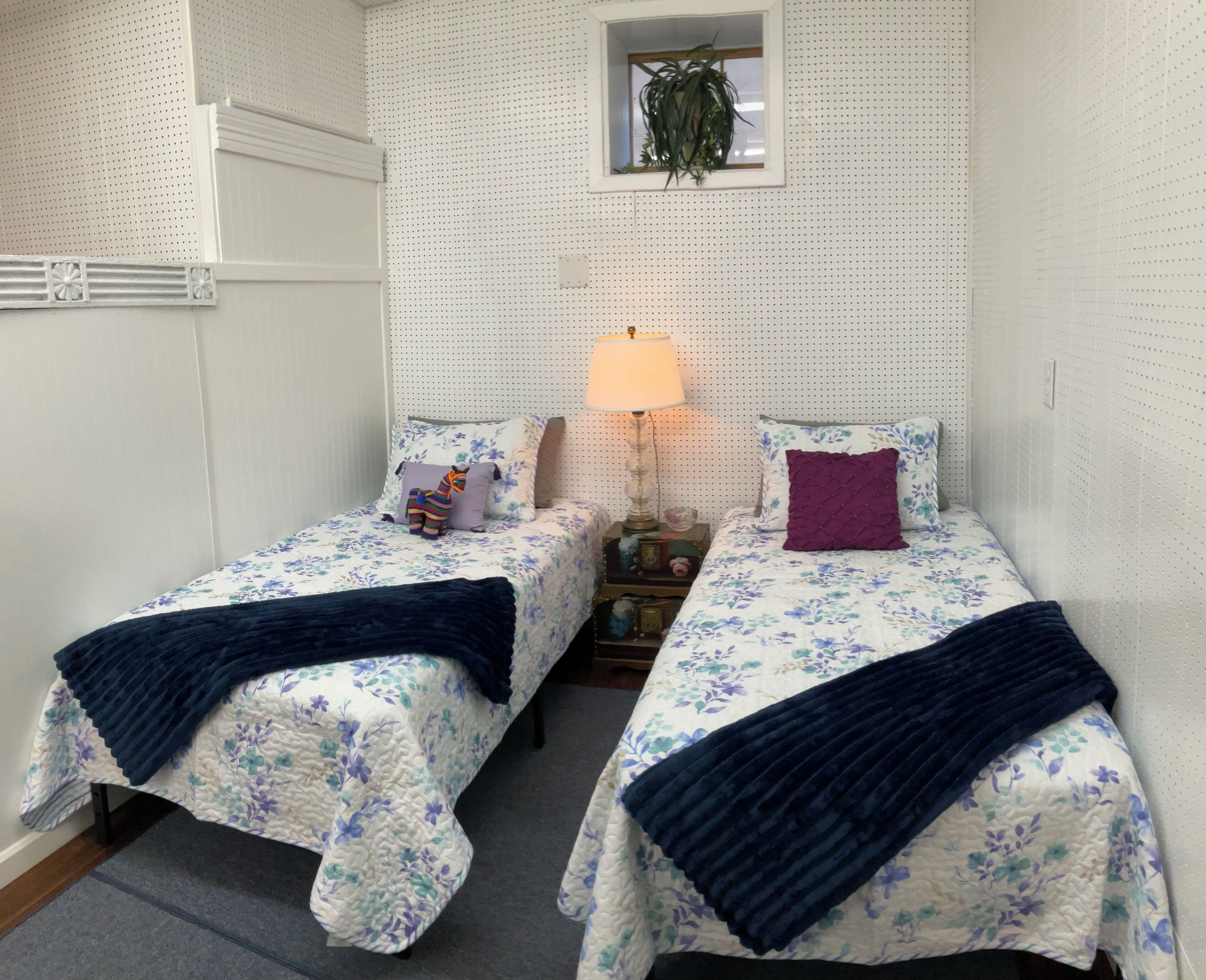 Et soveværelse med to senge, et natbord, en lampe og et lille vindue med en plante.