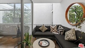 Uma sala de estar moderna com um sofá de cor escura, vários almofados estampados, um espelho redondo e um vaso com planta.