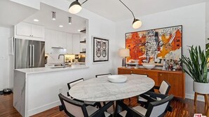 Cozinha moderna com armários brancos, geladeira de aço inoxidável e ilha de mármore. Há um espaço de jantar com mesa redonda e cadeiras, e um grande quadro abstrato na parede.